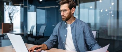 Homme barbu avec lunettes travaillant sur un ordinateur portable dans un bureau moderne, tenant une feuille de papier et utilisant une calculatrice