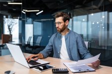 Homme barbu avec lunettes travaillant sur un ordinateur portable dans un bureau moderne, tenant une feuille de papier et utilisant une calculatrice